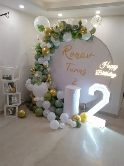 A sophisticated jungle-themed second birthday. This setup features a sage green and gold balloon arch, a personalized backdrop, a neon sign, and 'BABY' blocks.