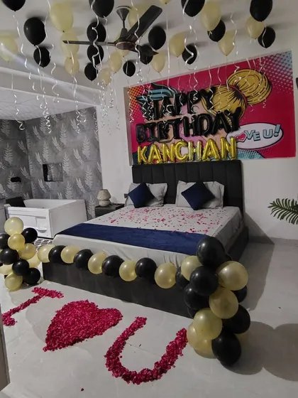 A personalized birthday surprise combining romance and celebration. This bedroom setup includes 'Happy Birthday' balloons with the person's name, rose petals on the floor spelling 'I Luv U', and a modern black and gold balloon theme.