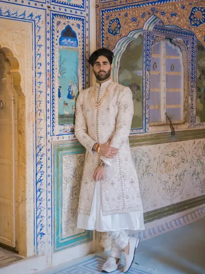 A groom's portrait that is both regal and composed. Posed against intricate blue and white palace tilework, his elegant sherwani and calm demeanor create a timeless and striking image.