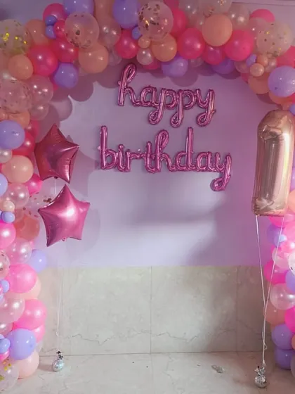 A classic first birthday balloon arch in pink, purple, and peach. This decoration includes confetti balloons for sparkle and 'Happy Birthday' foil letters, creating a sweet and simple backdrop for a home party.