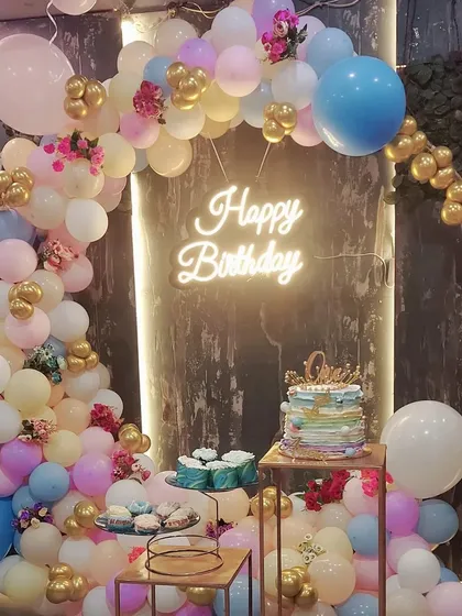A colorful and fun birthday party setup centered around a cake display. This design includes a pastel-colored organic balloon arch, a bright 'Happy Birthday' neon sign, and multi-level stands for the cake and desserts, all tied together with touches of gold and floral accents.