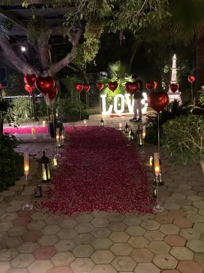 This expansive outdoor proposal scene showcases a long, dense path of red rose petals flanked by candles and red heart-shaped foil balloons. In the distance, our large, illuminated "LOVE" letters provide a stunning backdrop.