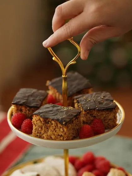 A hand reaching for a piece of chocolate-glazed plum cake from a tiered stand, adding a touch of action to the scene.