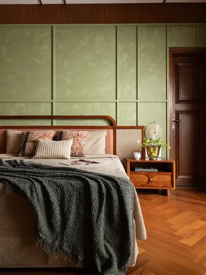 The master bedroom in our Indian heritage-inspired project. A sage green textured wall with paneling serves as a serene backdrop for the bed, which features a woven rattan and teak wood headboard. The herringbone wood flooring adds warmth and classic elegance.