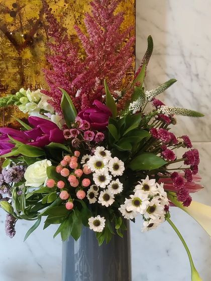 A contemporary floral arrangement in a sleek grey vase. The vibrant magenta tulips and pink astilbe pop against the delicate white daisies and greenery, perfect for a modern corporate setting.