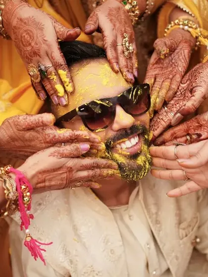 A close up of the groom's Haldi moment. Surrounded by the hands of his loved ones, his joyful smile shines through, even with sunglasses on.