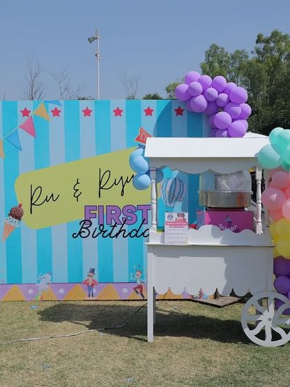 A sweet treats station at an outdoor twin's birthday carnival. The setup features a custom backdrop, a classic white cart for candy floss, and beautiful pastel balloon decorations.