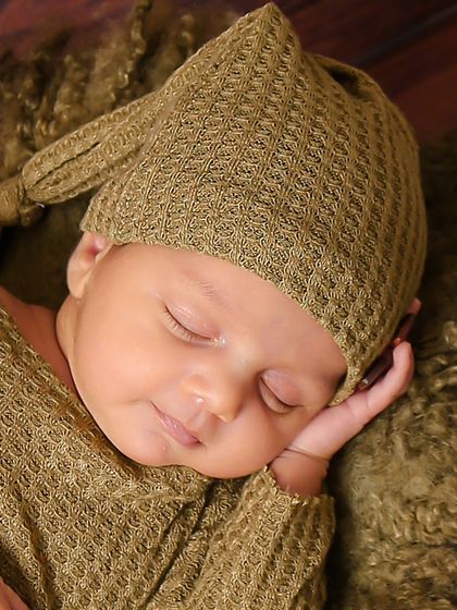 A sleeping baby in an olive green outfit and matching knotted hat. The hand-on-cheek pose is a classic that beautifully frames their peaceful face.