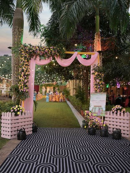 A charming entrance for a garden Haldi ceremony. The pink floral arch and patterned runner create a welcoming pathway, inviting guests into a space designed for celebration and togetherness.