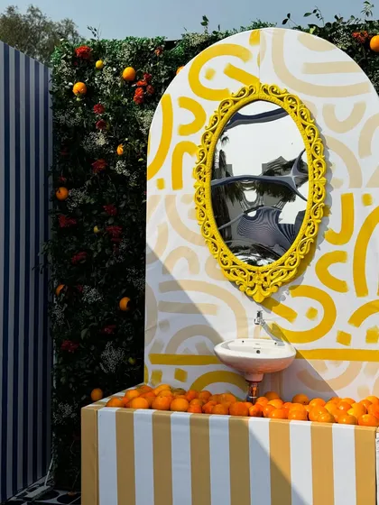 A whimsical and artistic corner from our Uttarayan setup. It features a vanity with a bright yellow ornate mirror, a sink filled with oranges, and a backdrop combining stripes with abstract patterns.
