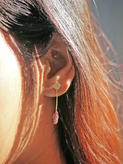 A close-up detail shot of a unique drop earring. The sunlight filtering through the model's hair highlights the raw texture of the pink gemstone, showing off the natural beauty of the material.