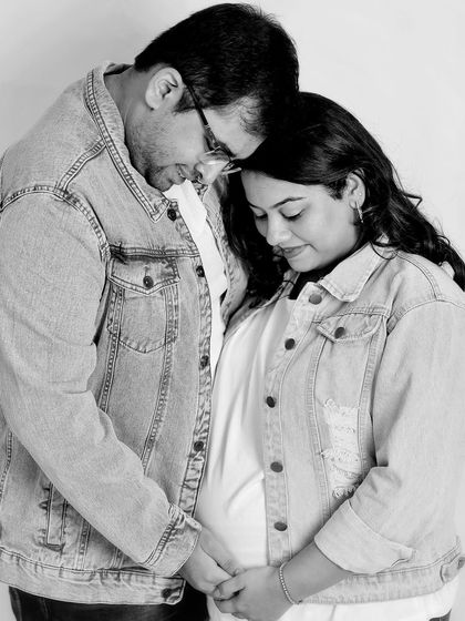 A casual and tender black and white photo. The couple, wearing matching denim jackets, shares a quiet moment, their heads together, focusing on the baby bump.
