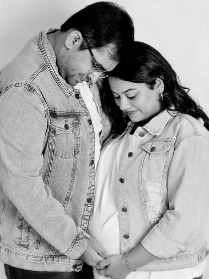 A casual and tender black and white photo. The couple, wearing matching denim jackets, shares a quiet moment, their heads together, focusing on the baby bump.