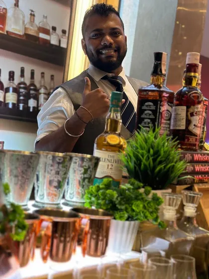 A happy bartender gives a thumbs-up from behind the whisky bar. This shot showcases the friendly and professional service I provide, along with the high-quality spirits and stylish copper mugs used for serving.