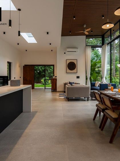 The open-plan living and dining area of a luxury villa. High ceilings with wooden paneling, skylights, and large glass walls create a bright, airy space that seamlessly connects with the lush green outdoors.