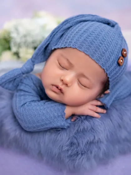 A close-up portrait of a sleeping newborn in a cozy blue hat. The soft purple fur underneath adds a touch of texture and color, highlighting the baby's peaceful expression.