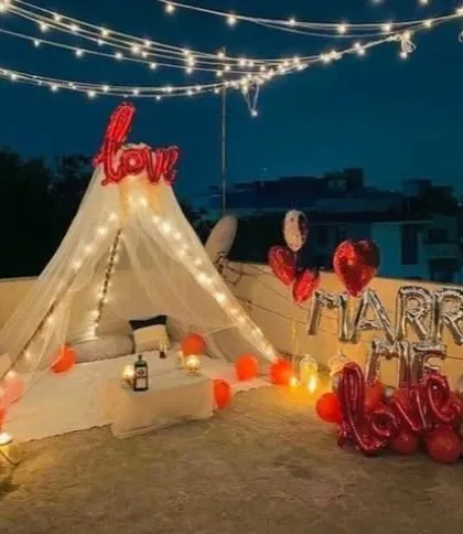 A dreamy rooftop proposal setup. A canopy tent adorned with fairy lights is set up under the night sky, with 'Marry Me' and 'Love' foil balloons making the intention clear.