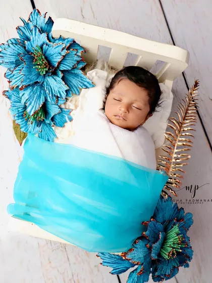 A baby sleeping on a white diwan, covered with a sheer blue blanket and surrounded by large blue flowers.