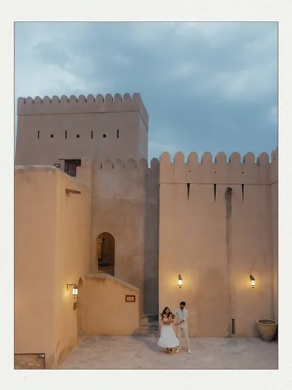 A couple dances in the courtyard of an Omani fort at dusk. The warm lights of the fort and the darkening blue sky create a magical and romantic atmosphere for this pre-wedding photo.