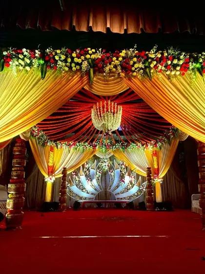 A full view of a traditional wedding mandap with rich yellow and red drapes. The floral work includes a thick border of red, yellow, and white flowers along the top and bouquets on the pillars, creating a warm and festive look.