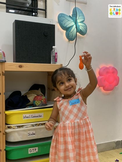 A smiling child holds up a small toy, her name tag visible. This highlights the personalized attention and joyful atmosphere we cultivate for each member.