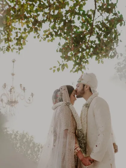 A dreamy, ethereal shot of a groom kissing his bride's forehead under a tree. The soft, hazy light and an out-of-focus chandelier create a magical and romantic wedding portrait.