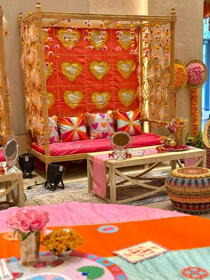A vibrant Mehendi seating area that blends tradition with playful details. The design features a canopy with a backdrop of heart-shaped mirrors, custom-printed cushions, and a fusion of Gujarati Patola and Rajasthani Gota work.