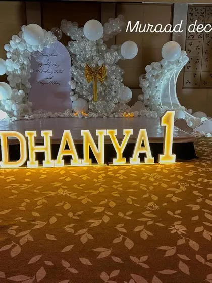A royal first birthday for a little princess. This grand setup features large, illuminated marquee letters spelling out her name on a patterned carpet, with an all-white, castle-themed backdrop.