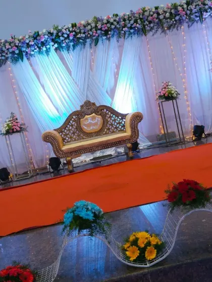 An angled shot of the wide reception stage, showing the orange carpet aisle and the decorative floral arrangements along the front. The blue uplighting gives the white drapes a cool, elegant tone.