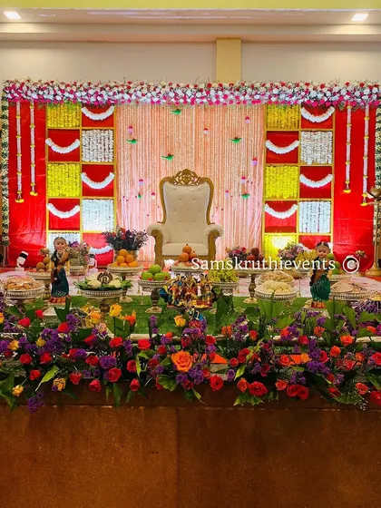 A wide shot of a classic red, yellow, and white themed baby shower stage. The foreground is filled with a bed of colorful flowers, adding a fresh and vibrant touch to the entire setup.