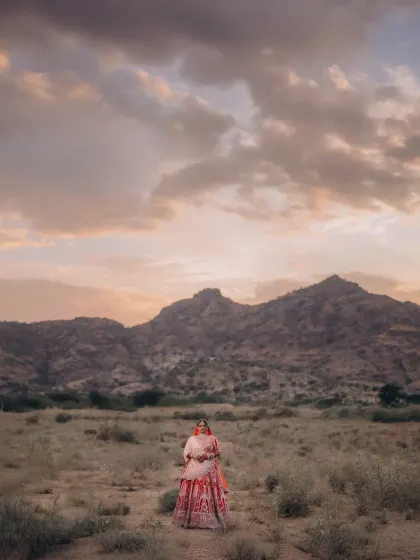 Nishi stands against a dramatic sky and mountain range. Her vibrant lehenga contrasts beautifully with the soft, dusky tones of the landscape at twilight.