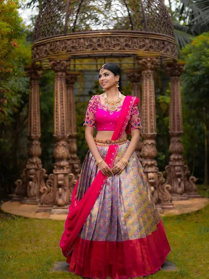 A full-length portrait showcasing a designer Kanjeevaram half saree. The grand architectural background adds to the regal feel of the outfit.