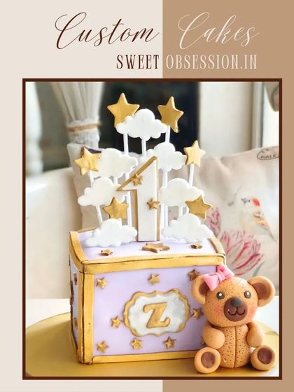 A whimsical first birthday cake designed as a gift box with a teddy bear, clouds, and stars.