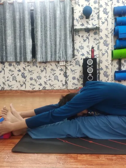 An instructor assists a student in a deep Paschimottanasana (Seated Forward Bend), using a yoga block for support. This hands-on guidance helps the student to safely explore their flexibility limits.