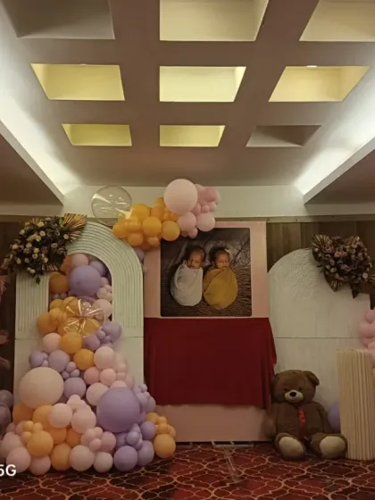 A lovely naming ceremony decoration for twins, featuring a photo of the babies at the center. The setup includes teddy bears, a picket fence, and pastel pink, purple, and orange balloons.