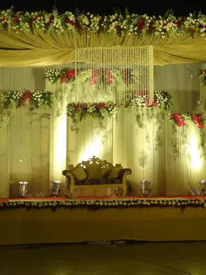 A classic stage decoration featuring a central crystal chandelier hanging from a draped ceiling. The backdrop is layered with fresh flowers in red and white, complemented by warm uplighting to create a grand and inviting focal point.