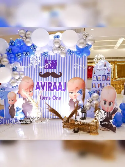 A playful photo booth for a Boss Baby party. The backdrop features a blue and white striped pattern with a mustache accent, surrounded by character cutouts and "BABY" blocks.