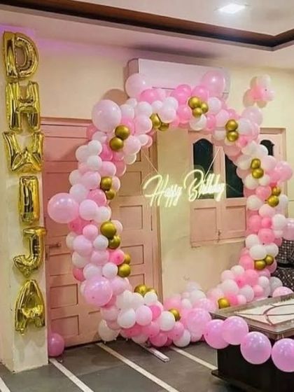 A pretty pink and gold balloon ring set up in a living room. The name 'DHVIJA' is spelled out vertically with large foil balloons next to the ring.