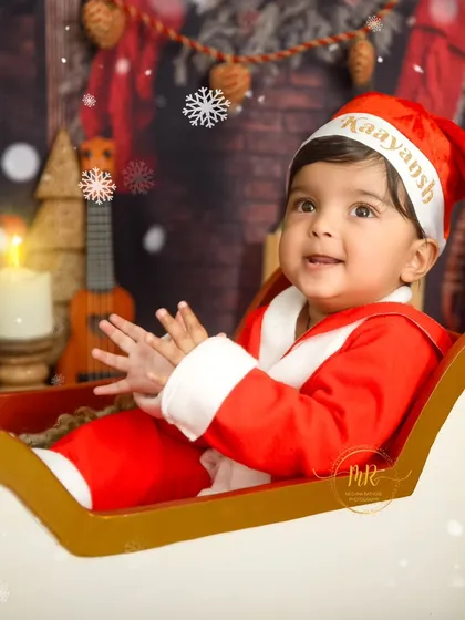 A happy baby wearing a personalized "Kaayansh" Santa hat, clapping his hands while sitting in a sleigh.