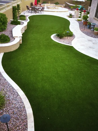 A free-form garden design using artificial turf to create flowing curves. The lawn is bordered by raised planters and a stone patio with a fire pit, showing the versatility of artificial grass.