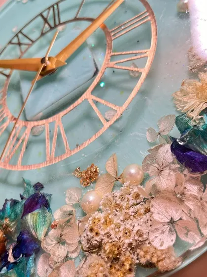 A close-up of a 12-inch wall clock with preserved garlands. The light blue resin base makes the white, blue, and purple flowers stand out, complemented by a classic Roman numeral dial.