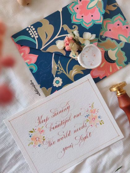 "Keep shining beautiful one, the world needs your light." A motivational card featuring calligraphy framed by delicate watercolor florals, sealed with wax on a beautiful patterned envelope.