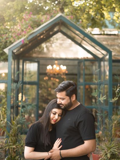 A tender and loving pre-wedding moment in a garden setting. The groom embraces the bride as she rests her head on his shoulder, creating a feeling of warmth and security.