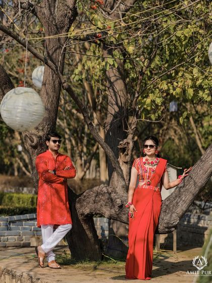 A stylish pose under a tree during their outdoor wedding festivities. The bride's modern red saree and the groom's classic kurta create a striking look against the rustic, natural background.