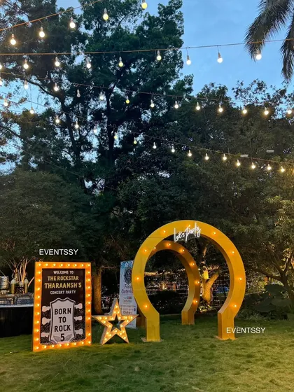 The rockstar party entrance at dusk, with string lights overhead creating a magical ambiance. The glowing marquee signs and arches stand out beautifully against the evening sky.