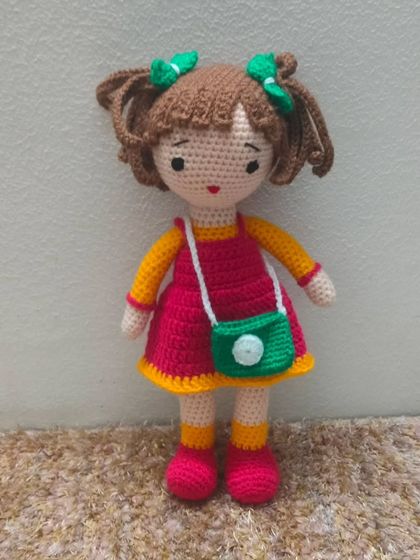 This cute doll has brown hair in pigtails and is wearing a red and yellow dress with a green bag.