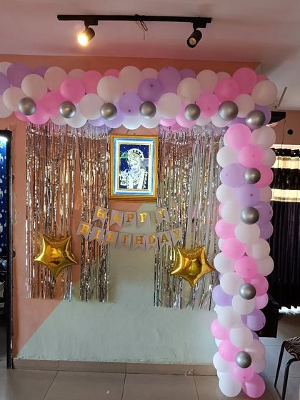 A pastel-themed birthday decoration with a pink, purple, and silver balloon arch against a tinsel curtain.