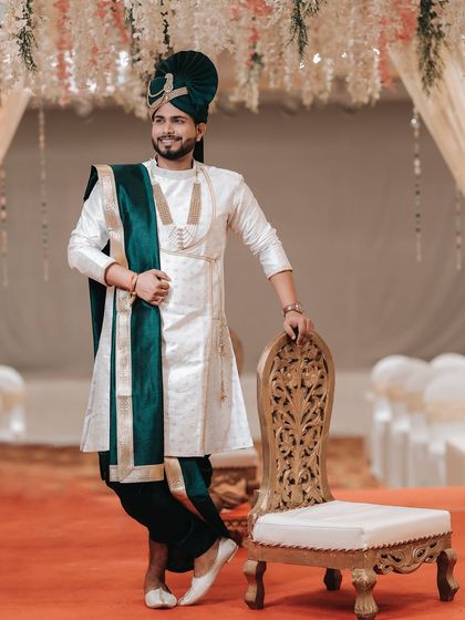 A stylish portrait of the groom in his elegant white and green sherwani. His confident pose next to the ornate chair makes for a regal wedding photo.