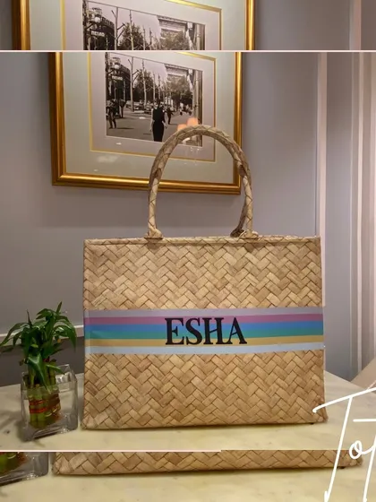 A natural straw tote bag with a pastel rainbow stripe and the name 'Esha'. This bag has major beach vacation vibes.