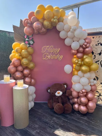 A bright and cheerful daytime birthday party setup on a terrace. This features a solid pink backdrop with a 'Happy Birthday' sign, framed by an organic balloon arch in gold, rose gold, and white, and accented with pedestals for the cake or gifts.